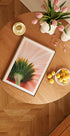 a wooden table with a framed picture of a flower, a bowl of fruit, and a vase of pink flowers.