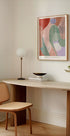 a minimalist workspace with a wooden desk, a woven chair, a lamp, and a framed abstract painting on the wall.