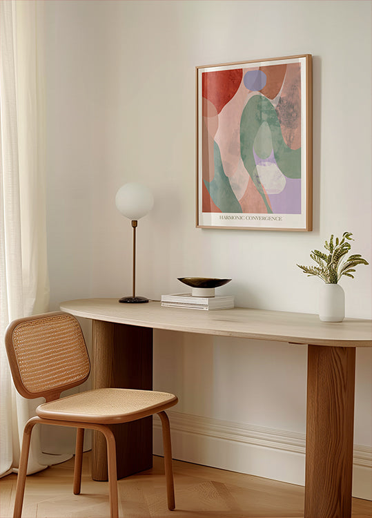 a minimalist workspace with a wooden desk, a woven chair, a lamp, and a framed abstract painting on the wall.
