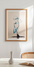 a framed artwork of a water droplet suspended in mid-air, with a blue and white color scheme. The artwork is displayed on a white wall above a table with a white vase and a book.